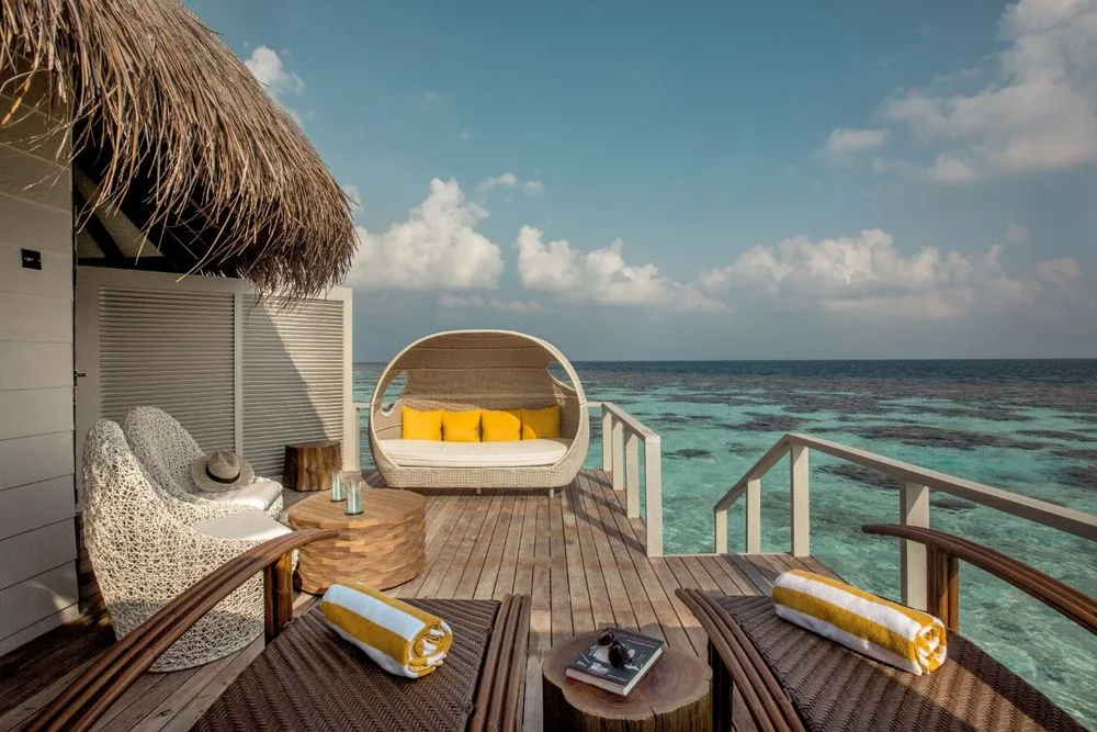 DRIFT Thelu Veliga Retreat, villa overwater, Alif Dhaal (South Ari) Atoll, Maldives, Water Villa deck under the thatched-roof overhang, woven-rattan daybed with the property-signature yellow accent pillows and matching towels, two open-weave white lounge chairs, steps descending into the turquoise lagoon.