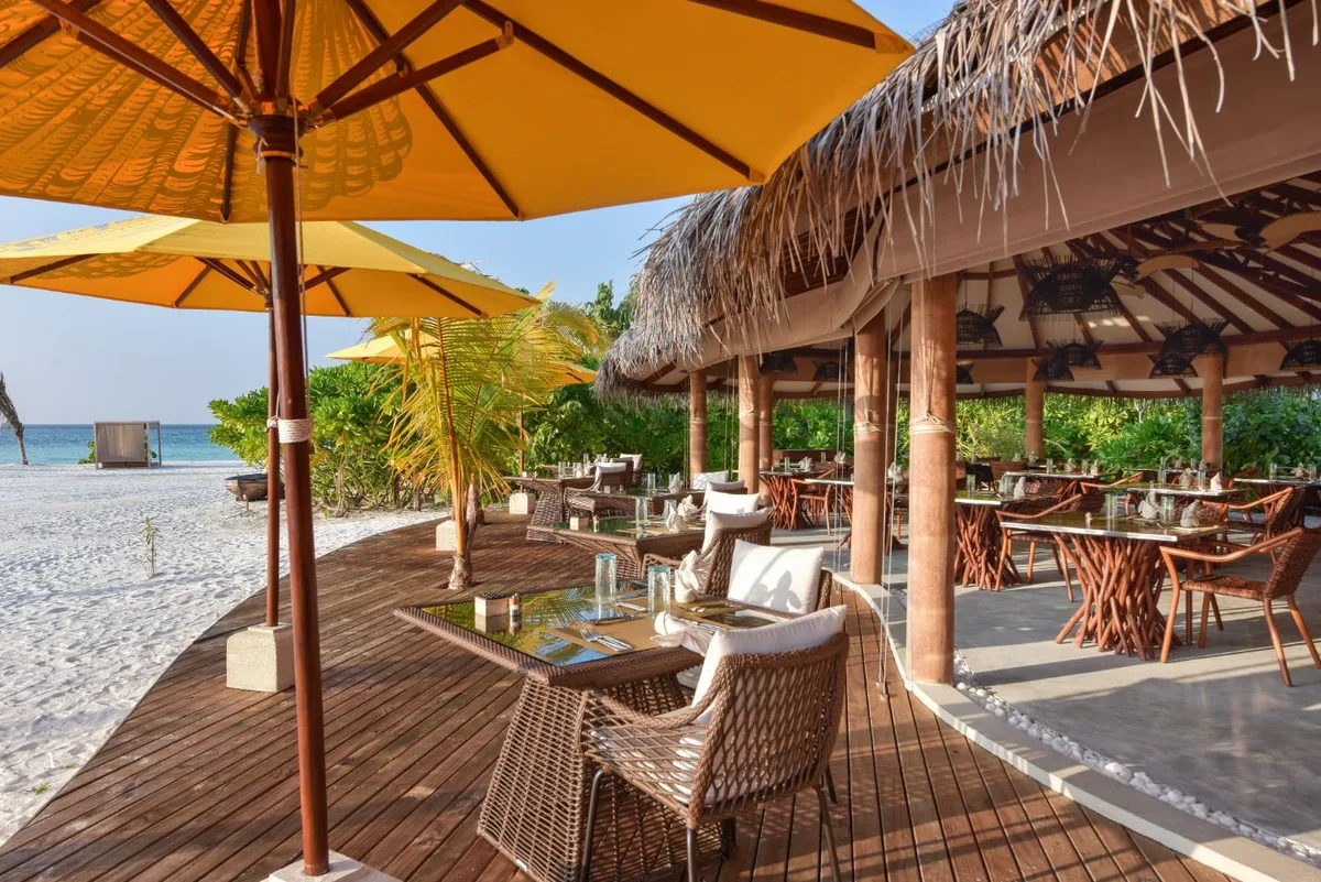 DRIFT Thelu Veliga Retreat, dining, Alif Dhaal (South Ari) Atoll, Maldives, Beachside restaurant pavilion, thatched-roof structure with rope-bound timber columns and the property-signature yellow canvas umbrellas over woven dining chairs at glass-topped tables on the wooden deck, beach extending to the lagoon beyond.