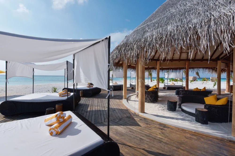 DRIFT Thelu Veliga Retreat, dining, Alif Dhaal (South Ari) Atoll, Maldives, Beach bar and lounge cluster, thatched-roof bar pavilion with dark-woven banquette sofas and yellow accent pillows inside, white canvas-shade pods over double-bed cabanas with rolled yellow towels on the deck.