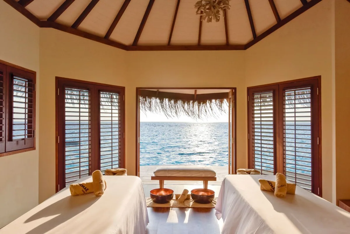 DRIFT Thelu Veliga Retreat, spa, Alif Dhaal (South Ari) Atoll, Maldives, Spa couples treatment room with warm-yellow walls and a dark-wood pitched-rafter ceiling, twin massage beds with MAEMAE-branded folded yellow-towel rolls, wooden plantation shutters on either side, open French doors framing the thatched-roof eaves and the lagoon horizon.