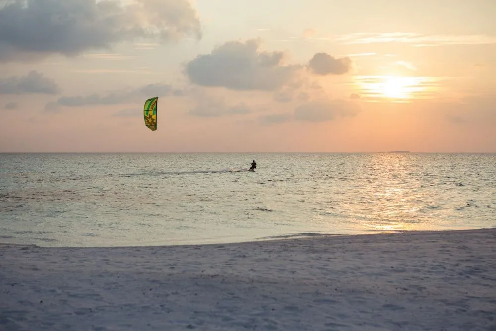 DRIFT Thelu Veliga Retreat, activities, Alif Dhaal (South Ari) Atoll, Maldives, Kitesurfer at sunset in the silver lagoon under a green-and-yellow kite (property-palette signature), figure silhouetted against the pink-orange horizon, white-sand foreground. The boutique adults-only operational tone calibrated for the watersports-and-relaxation pairing.
