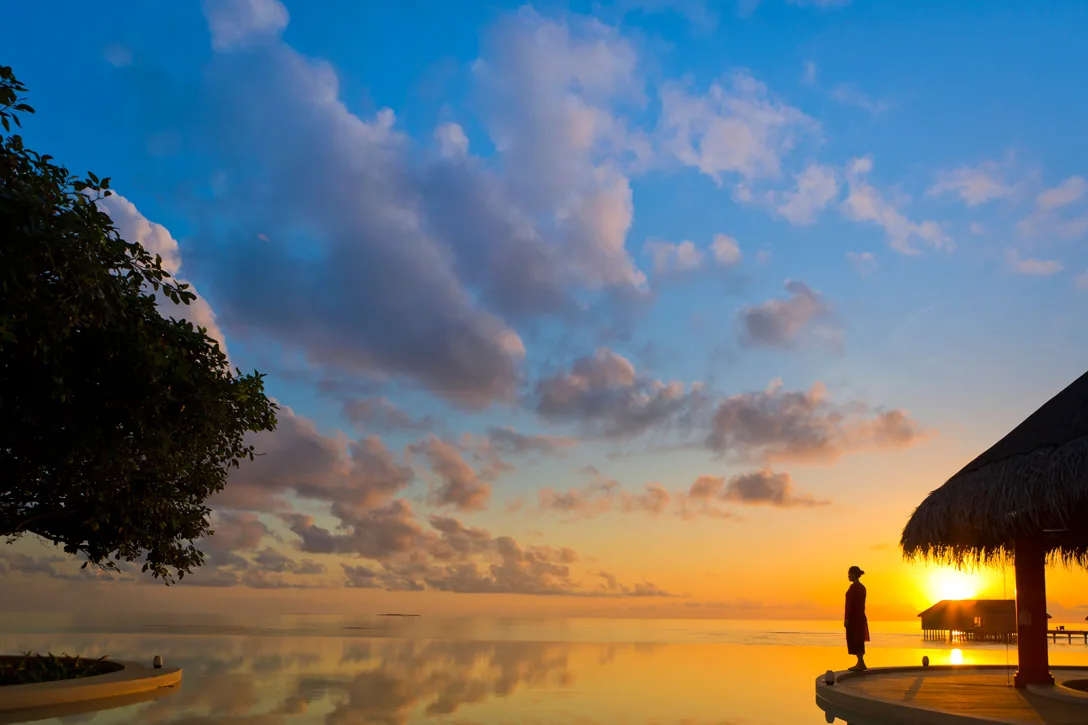 Dusit Thani Maldives, setting, Baa Atoll, Maldives, Infinity pool deck at sunset, the silhouetted guest standing at the pool edge with the thatched-eave bar pavilion at the right frame edge, dramatic cloud-banked sky reflected on the glass-still water surface.