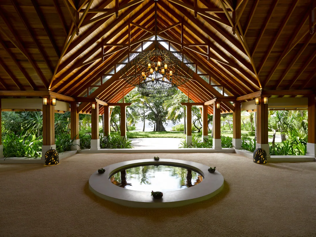 Dusit Thani Maldives, setting, Baa Atoll, Maldives, Arrival lobby pavilion interior, the soaring timber-truss thatched-cathedral ceiling with the woven-vine pendant chandelier, central round reflection pond with carved-stone turtles on the rim and the bodhi-tree silhouette visible through the open archway.