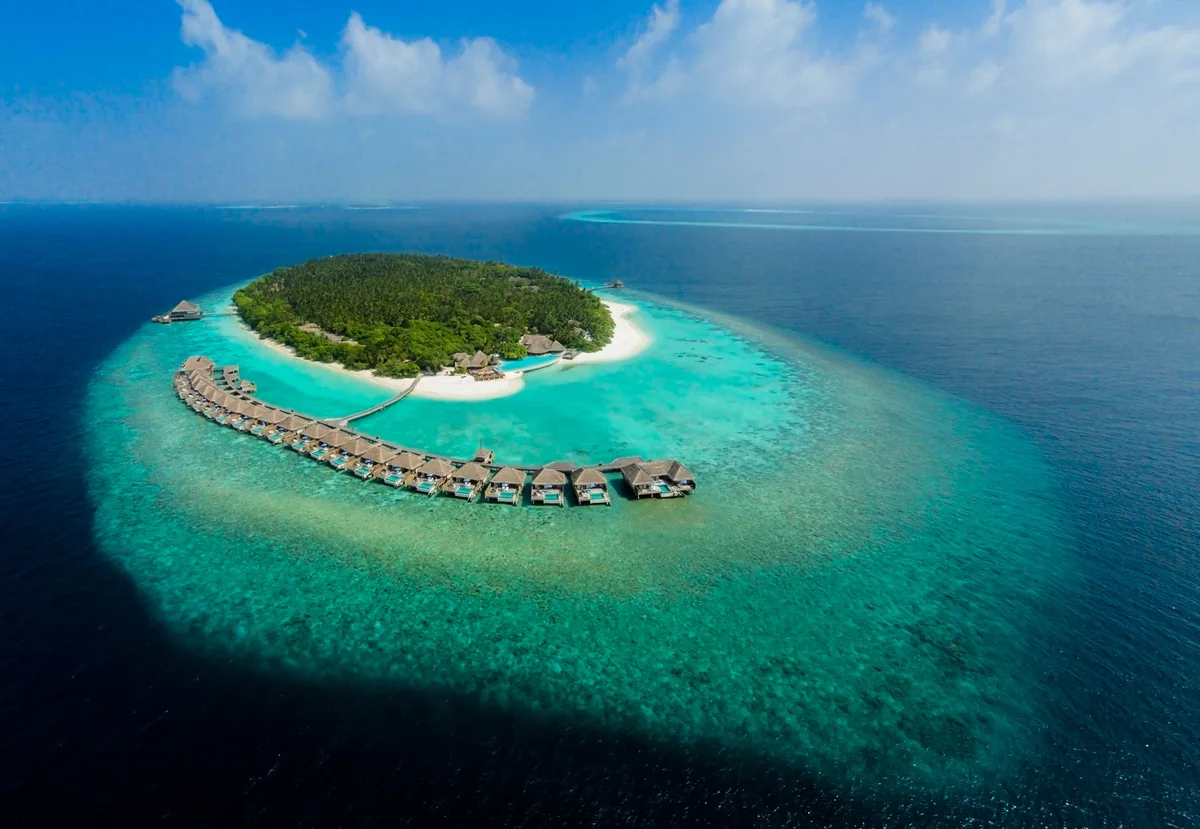 Dusit Thani Maldives, aerial, Baa Atoll, Maldives, Mudhdhoo island from the drone height, the oval coral reef and the signature curved row of thatched-pyramid overwater villas tracing the southeast lagoon edge, dense jungle interior with the central pool deck visible and the white-sand beach point.