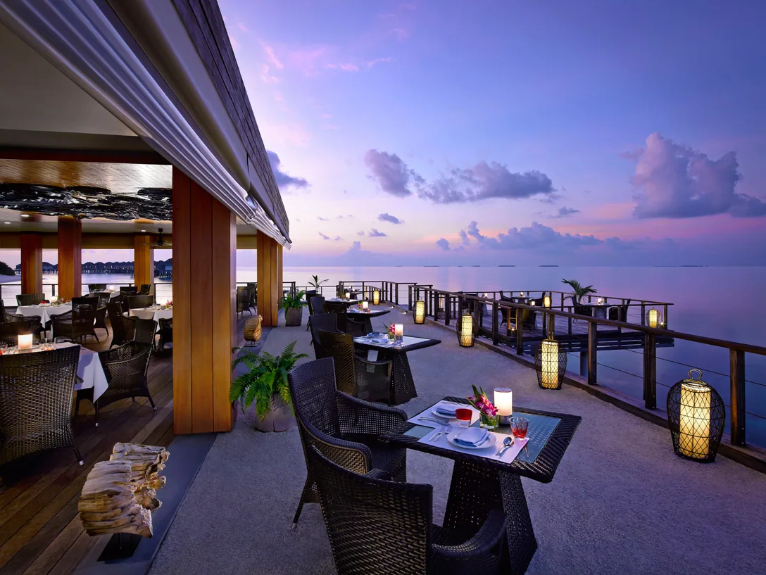 Dusit Thani Maldives, dining, Baa Atoll, Maldives, Benjarong Royal Thai restaurant at twilight, the open overwater terrace with woven-rattan armchairs, white-linen tables lit by glass-cylinder candles inside basket-woven lanterns, the lagoon stretching to the pink-and-purple horizon.