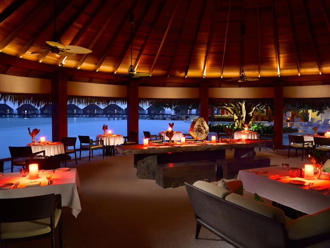 Dusit Thani Maldives, dining, Baa Atoll, Maldives, Sea Grill interior at night, the soaring timber-frame thatched roof glowing warm above white-linen tables, the central communal stone-base table with a golden bowl centrepiece and the overwater villa row visible through the open-eave at the back.