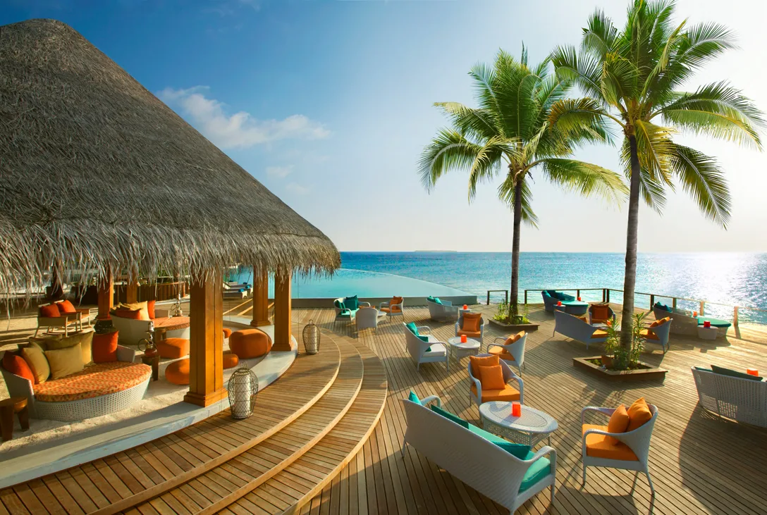 Dusit Thani Maldives, dining, Baa Atoll, Maldives, Sand Bar deck, the open thatched-eave bar with orange-and-teal cushioned rattan seating on hardwood decking, the infinity edge merging the deck into the lagoon and tall coconut palms framing the horizon.