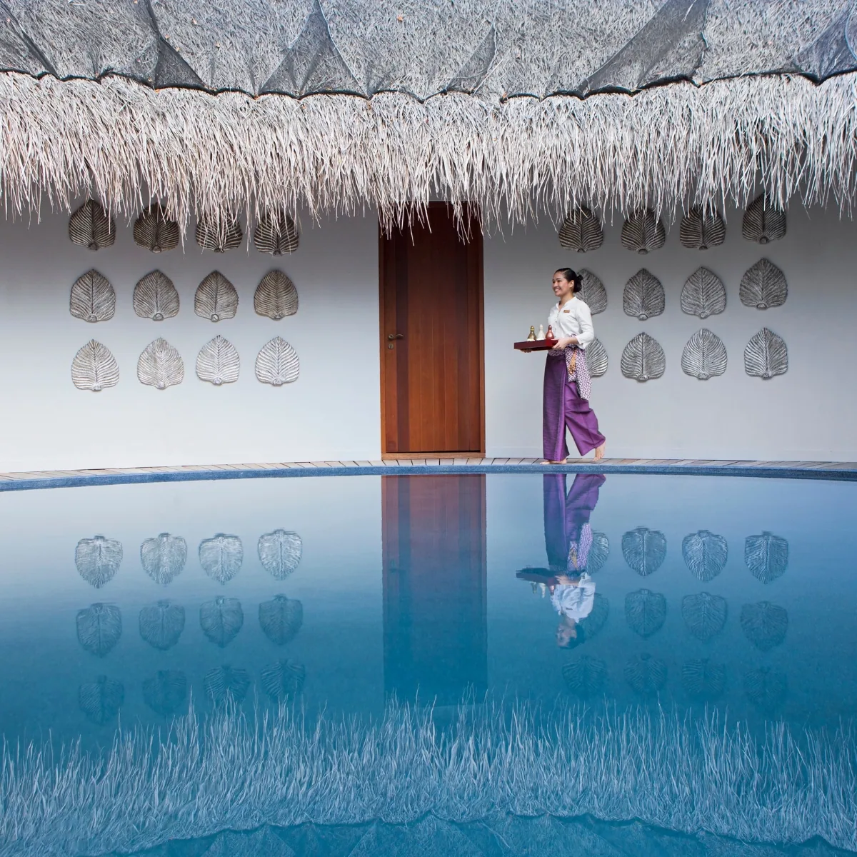 Dusit Thani Maldives, spa, Baa Atoll, Maldives, Devarana Spa pavilion exterior, the thatched-fringe roof over the white wall stencilled with the silver bodhi-leaf botanical motif, dark teak door and the spa therapist in white blouse with purple Thai-cut skirt carrying a treatment tray along the reflection-pool edge.