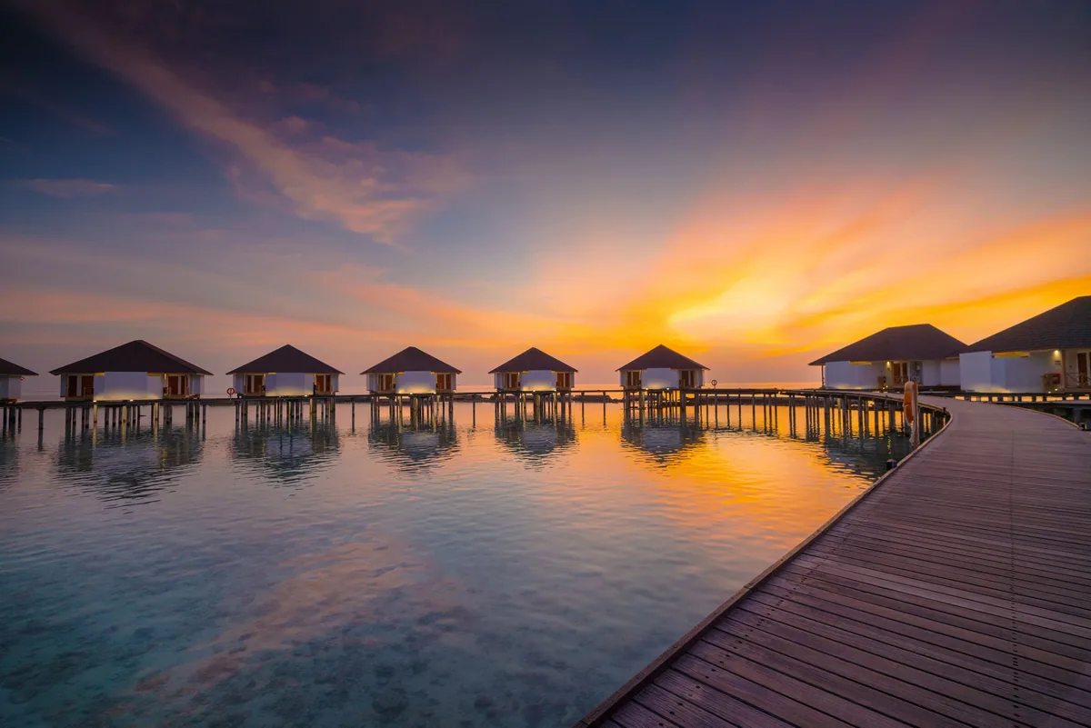 Cinnamon Ellaidhoo Maldives, lifestyle, Alif Alif Atoll (North Ari), Maldives, Water Bungalow row at sunset from the boardwalk perspective, the pyramid-roof villas silhouetted against a fiery pink-orange sky, the wooden boardwalk extending into the right of the frame, the OWV reflection in the still lagoon.