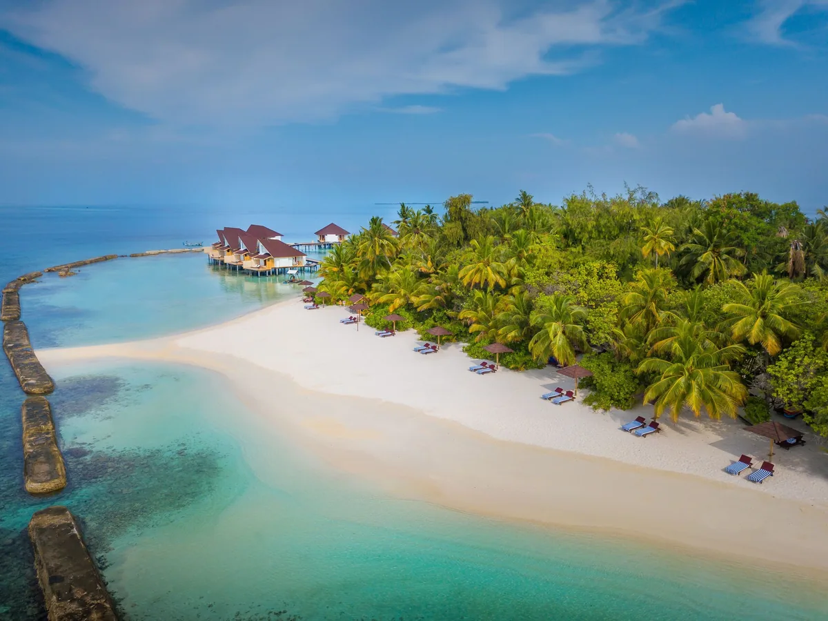 Cinnamon Ellaidhoo Maldives, setting, Alif Alif Atoll (North Ari), Maldives, House-reef-edge beach aerial, the sand line dotted with sun-loungers and red umbrellas, brown-tile pyramid-roof Water Bungalow row extending into the lagoon at the left, and the reef-protection wall arcing in the foreground (the 30-metre-from-beach reef edge that gives Ellaidhoo its shore-access dive identity).