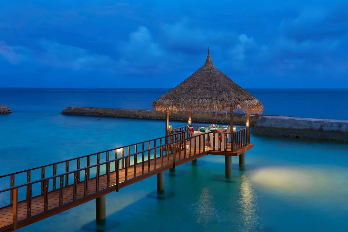 Cinnamon Ellaidhoo Maldives, dining, Alif Alif Atoll (North Ari), Maldives, Overwater private dining pavilion at twilight, octagonal thatched roof at the end of a wooden walkway over the lagoon, the table set for two inside the pavilion with candles and lanterns, the calm lagoon reflecting the blue evening sky.