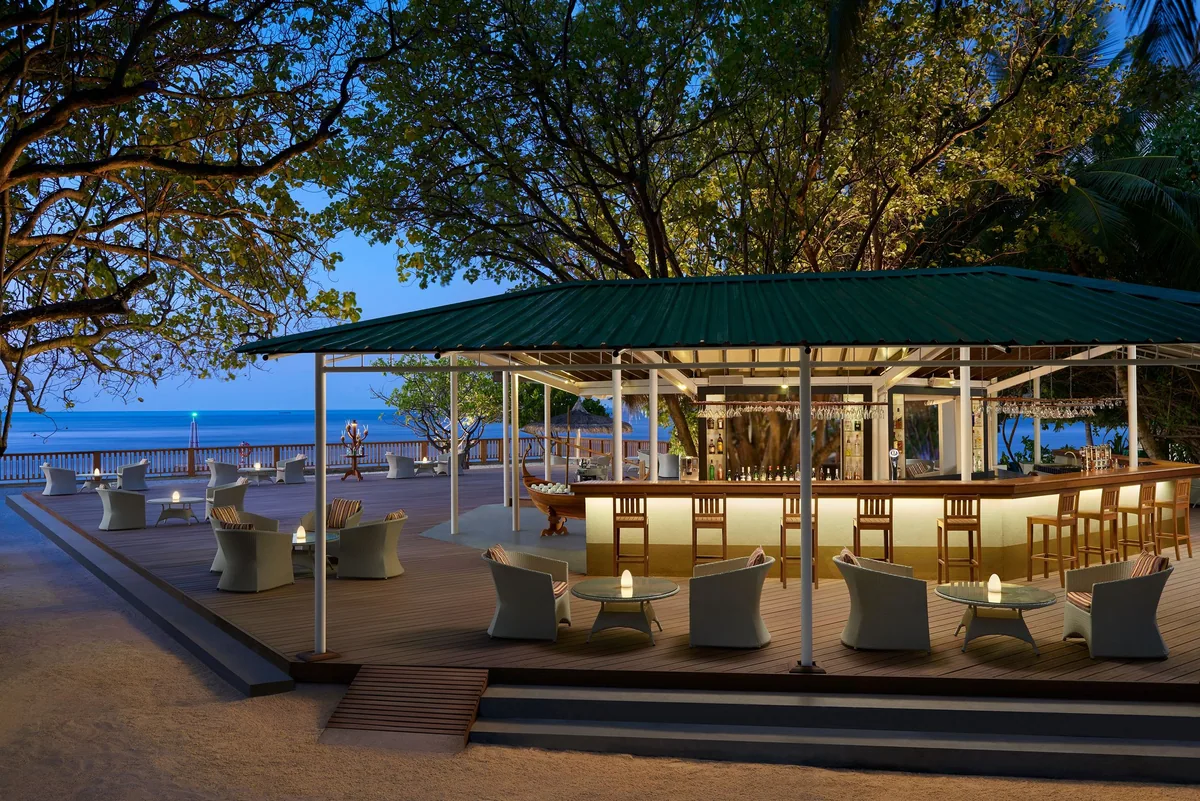 Cinnamon Ellaidhoo Maldives, dining, Alif Alif Atoll (North Ari), Maldives, Iroushenee Bar at dusk, open-air pavilion under a green-canopy roof with a lit bar counter and stools, tables and chairs arranged around the deck perimeter, tropical foliage framing the entry and the ocean horizon catching the last light.