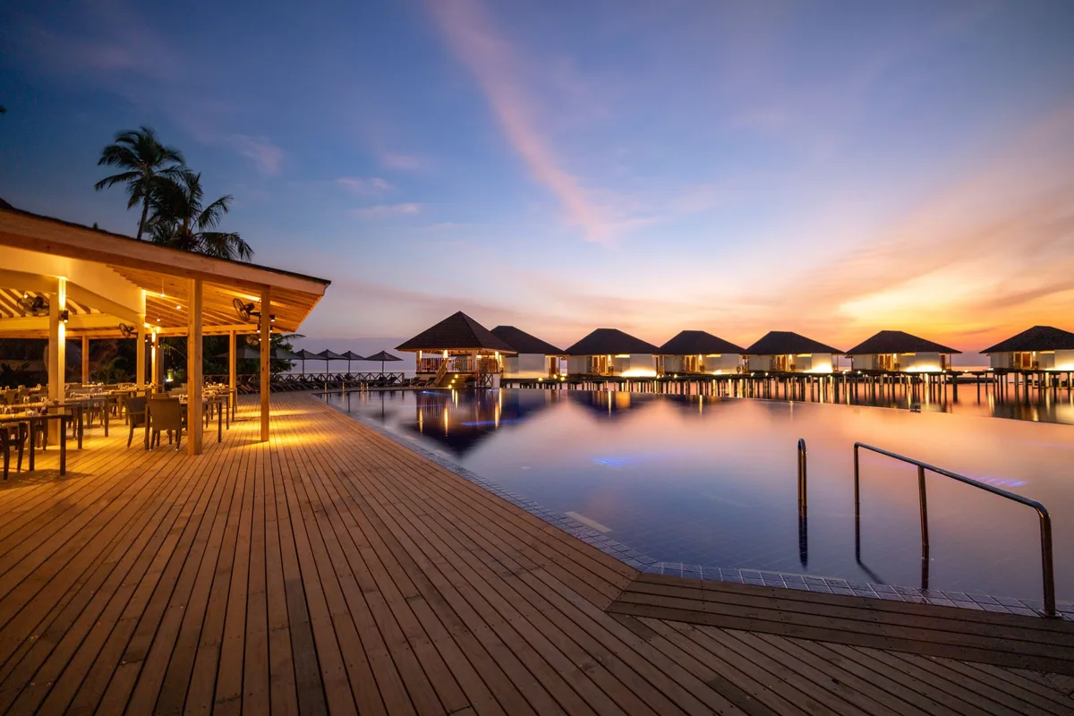 Cinnamon Ellaidhoo Maldives, dining, Alif Alif Atoll (North Ari), Maldives, Malamathi Restaurant pool deck at sunset, the infinity pool reflecting the brown-tile pyramid-roof Water Bungalow row, the open-side restaurant pavilion lit from within at the left, the dramatic pink-orange-yellow sunset sky transitioning to violet.