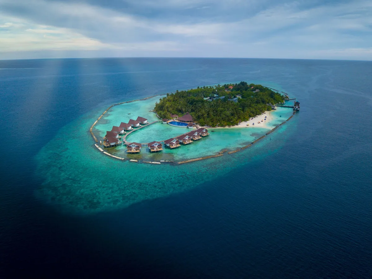 Cinnamon Ellaidhoo Maldives, setting, Alif Alif Atoll (North Ari), Maldives, Full Ellaidhoo island aerial, oval reef-protected lagoon with the brown-tile pyramid-roof Water Bungalow row arcing along the southern lagoon edge, palm-covered main island and reef edge running the full perimeter. The structural reef-protected island geometry as the property's setting identity.