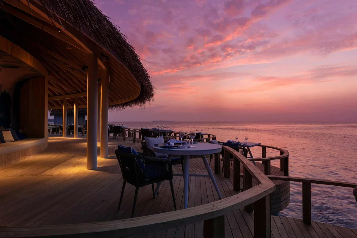 Emerald Faarufushi, Mediterraneo interior deck at sunset with curved wooden deck, navy rope-back chairs at round tables set with wine glasses, thatched eave and pink sunset lagoon, Raa Atoll, Maldives