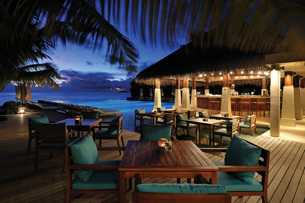 Emerald Faarufushi, Carnivorous South American grill at twilight with teal-cushion wooden chairs, square tables, central thatched bar with pendant lights, pool and palms, Raa Atoll, Maldives