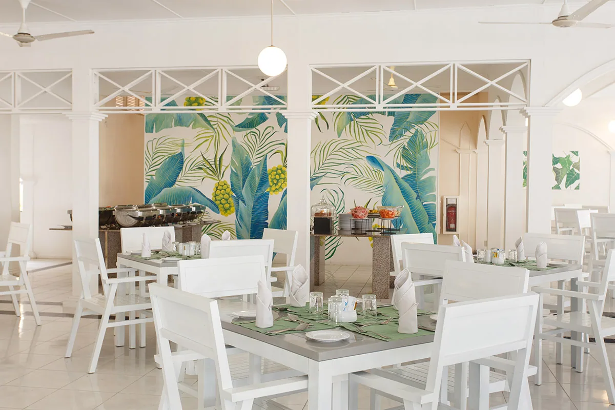 Equator Village, dining, Seenu (Addu) Atoll, Maldives, Principal all-day buffet pavilion: banana-leaf print feature wall behind the chafing-dish buffet line, globe pendant lights and ceiling fans overhead, white-painted timber four-tops with paper napkin tents on green table-runners, white cross-truss roof structure.