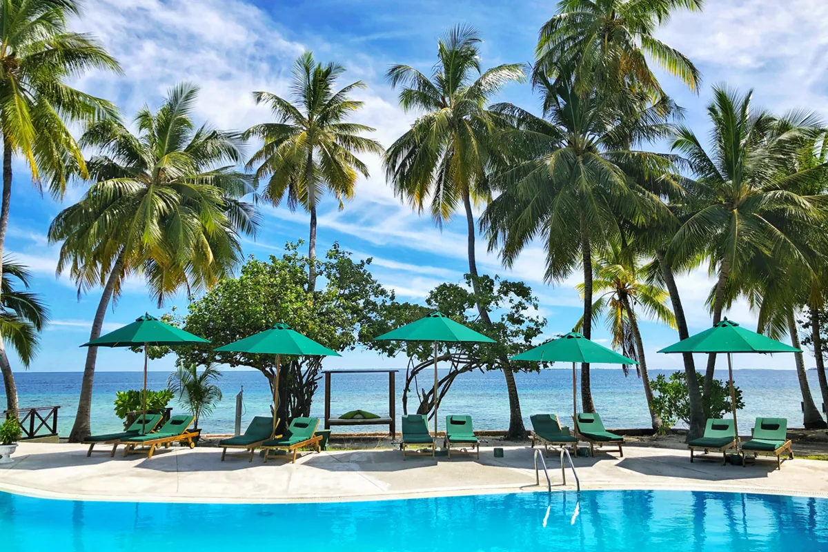 Equator Village, setting, Seenu (Addu) Atoll, Maldives, Pool-front setting with five teal-green parasols in a row over teal-cushioned sun loungers, palm canopy framing both sides, the lagoon visible directly beyond the pool deck, the resort's signature teal palette repeating across umbrellas + cushions + matching wicker mats.