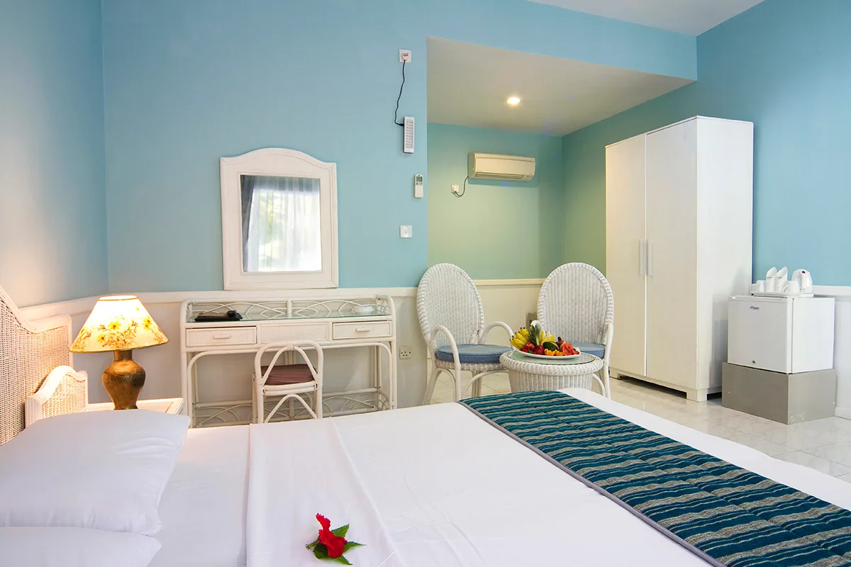 Equator Village, villa beach, Seenu (Addu) Atoll, Maldives, Standard Bungalow interior: teal-washed walls above white wainscoting, white wicker writing desk and tub chairs, hibiscus laid on the made bed with a teal-striped runner, welcome fruit platter on the rattan side table, minibar and mounted air-conditioning unit visible.