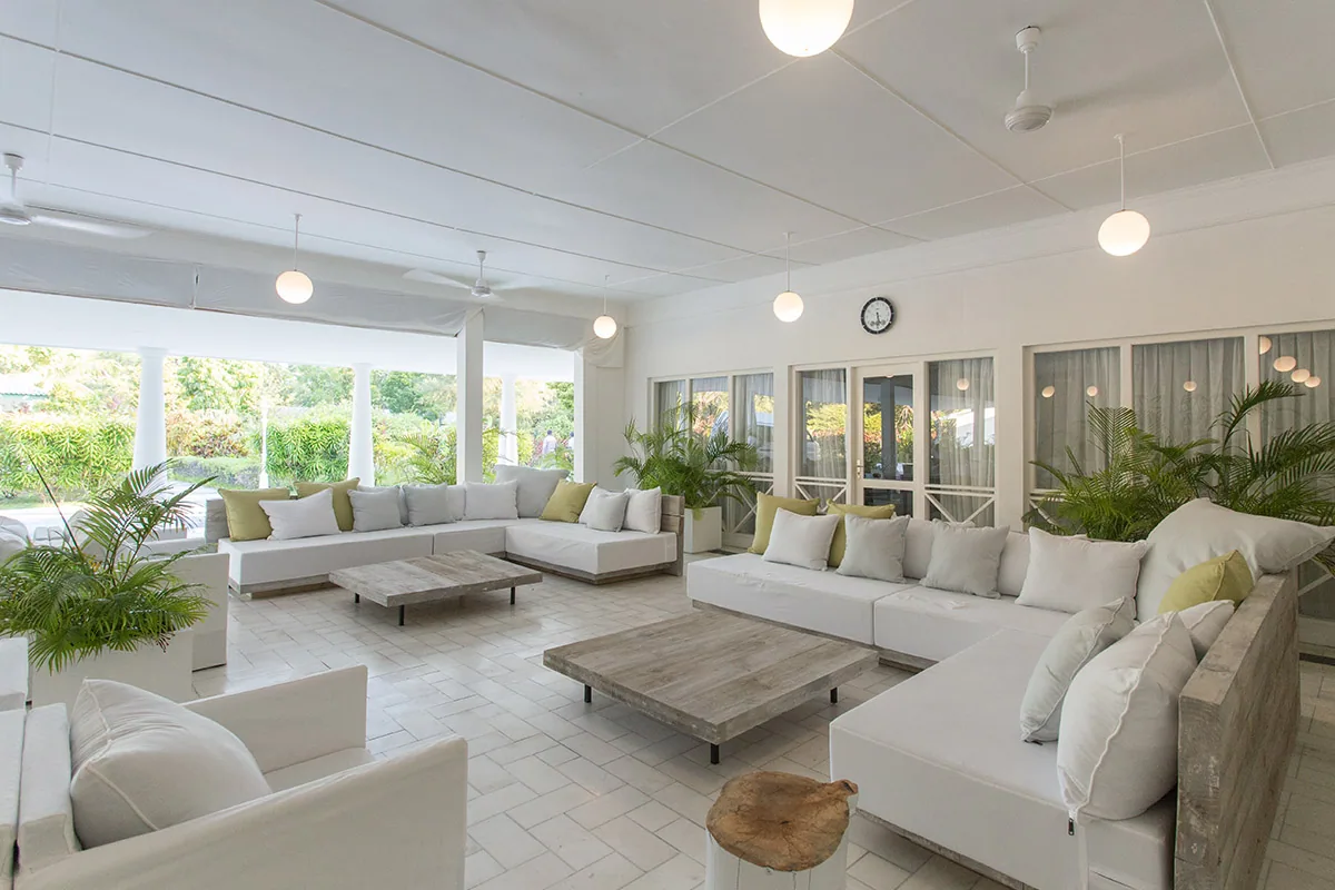 Equator Village, setting, Seenu (Addu) Atoll, Maldives, Open-sided main lounge with twin L-shaped white sofas facing reclaimed-timber low coffee tables, chartreuse cushion accents repeating across the seating, globe pendant lights and ceiling fans, framed garden view through the open colonnade.