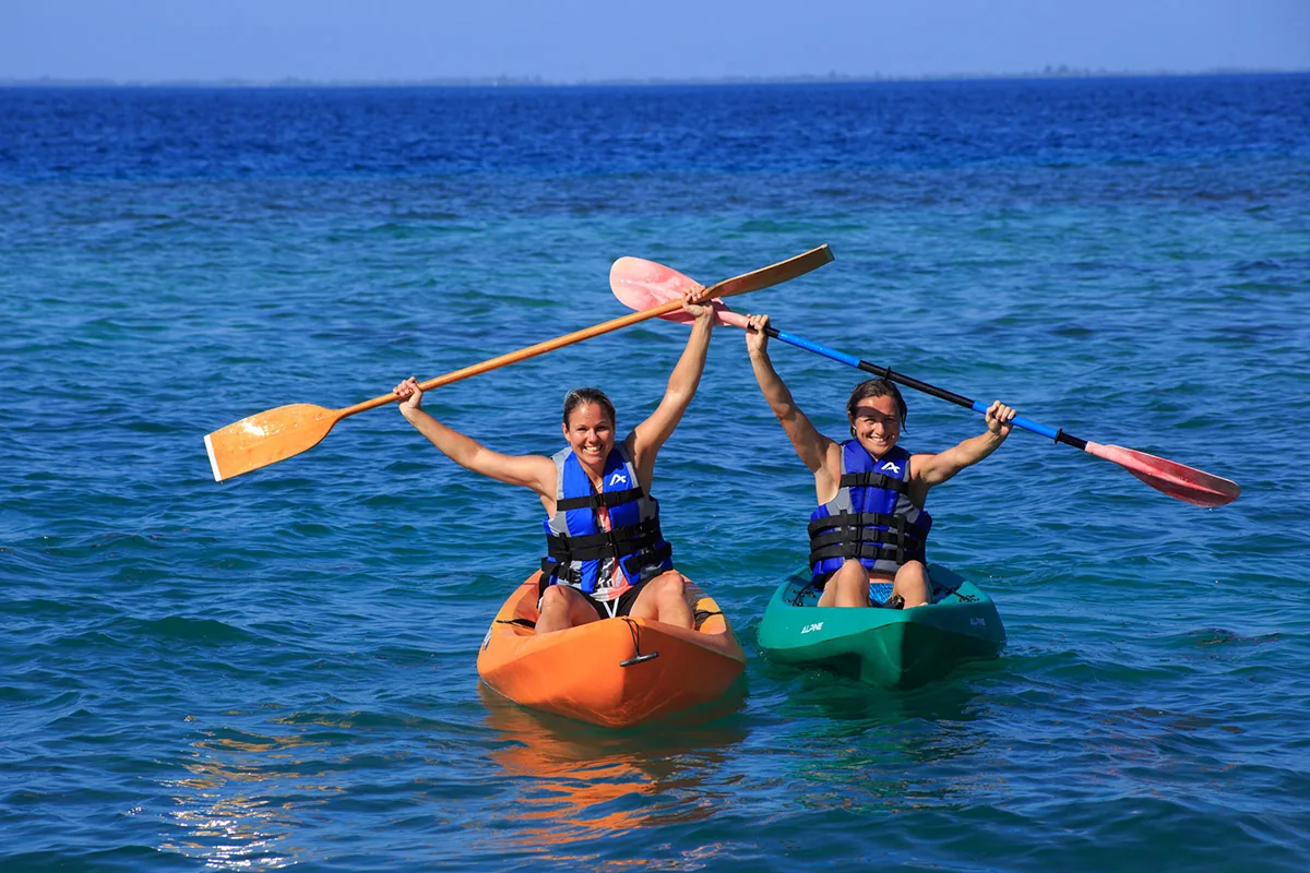 Equator Village, diving, Seenu (Addu) Atoll, Maldives, Twin guests in life jackets paddling tandem sit-on-top kayaks (orange + teal) on the calm Addu lagoon, deep-blue open water and reef line visible at the horizon; the equator-line dive-cluster lagoon doubles as the watersports launching ground.