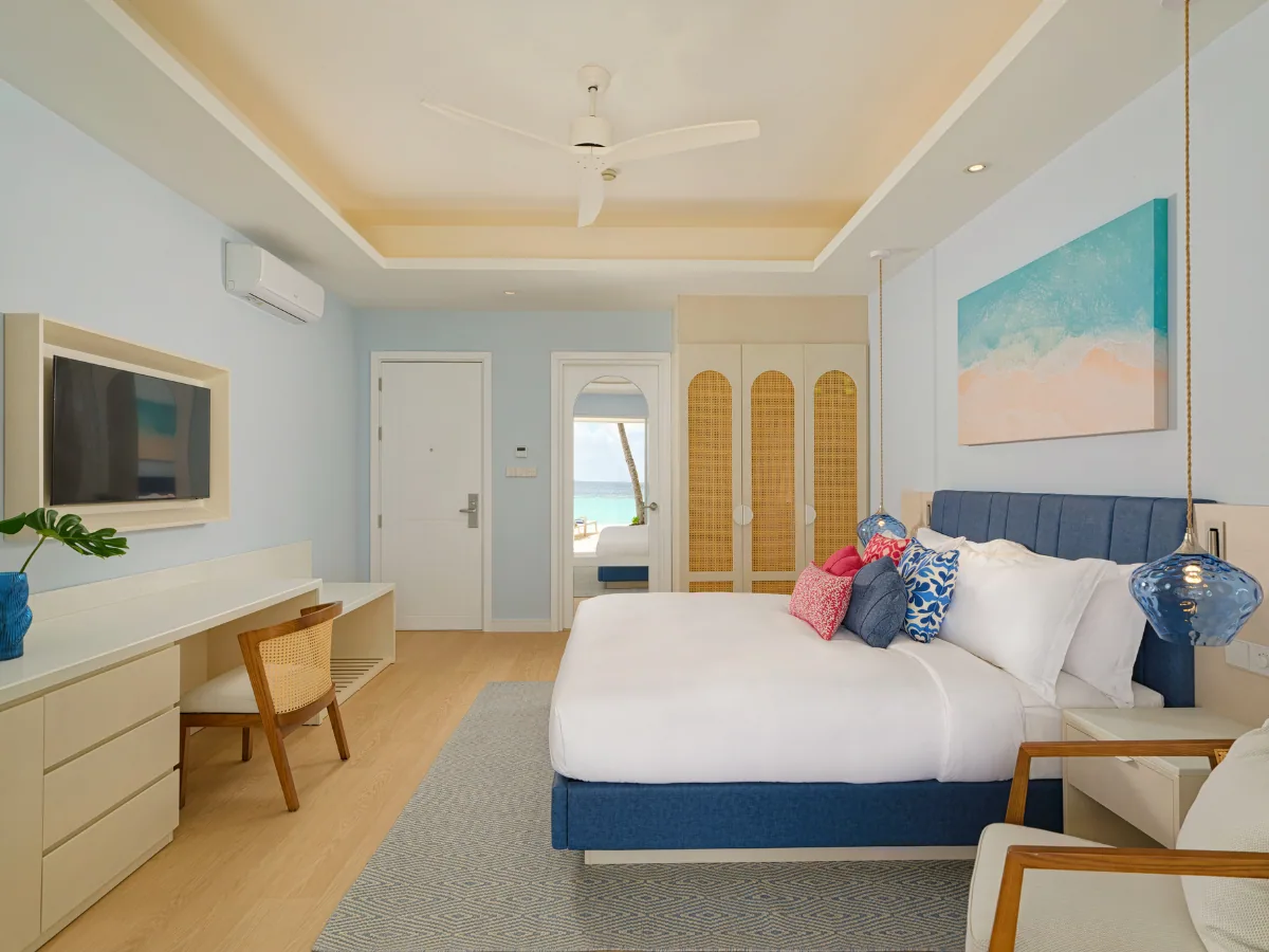 Eri Maldives, villa beach, North Malé Atoll, Maldives, Refreshed villa interior (Eri Maldives rebrand): soft blue-and-white palette with a king bed under a deep-blue tufted upholstered headboard, multicolour scatter cushions (turquoise, orange, pink, pattern-print), blue-print canvas art panel above the bed, white tray ceiling with ceiling fan, navy-louvre door at the side, light walnut floor.