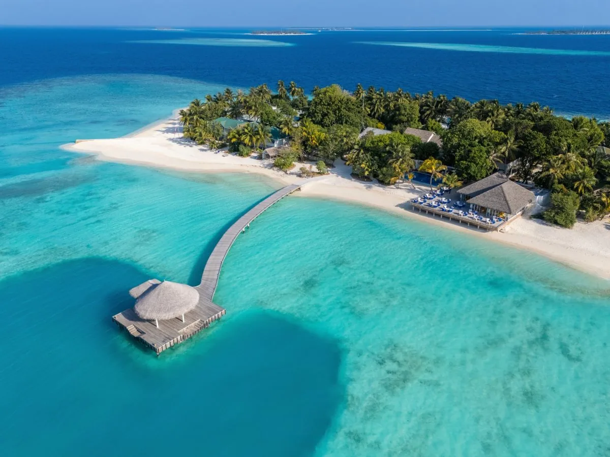 Eri Maldives, aerial, North Malé Atoll, Maldives, Eriyadu oblique aerial alt-angle: white sand beach perimeter with the thatched over-water Aabaru pavilion at the end of a curved jetty extending into the lagoon, the beachside thatched Beach Shack pavilion with sun loungers visible at the right edge, the vibrant turquoise lagoon with darker reef patches, the deep blue ocean and neighbouring atoll on the horizon.