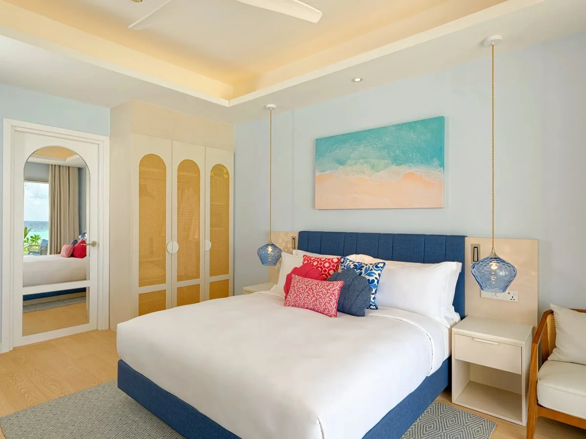 Eri Maldives, villa beach, North Malé Atoll, Maldives, Premium Beach Villa interior: wider spatial composition with a king bed under a deep-blue tufted headboard, twin hanging blue-glass pendant lights, a watercolour beach-aerial canvas mounted above the headboard, an arched wardrobe with rattan-cane louvre panels at the side, ornate floor mirror reflecting another bedroom view, ceiling fan above, light parquet floor.