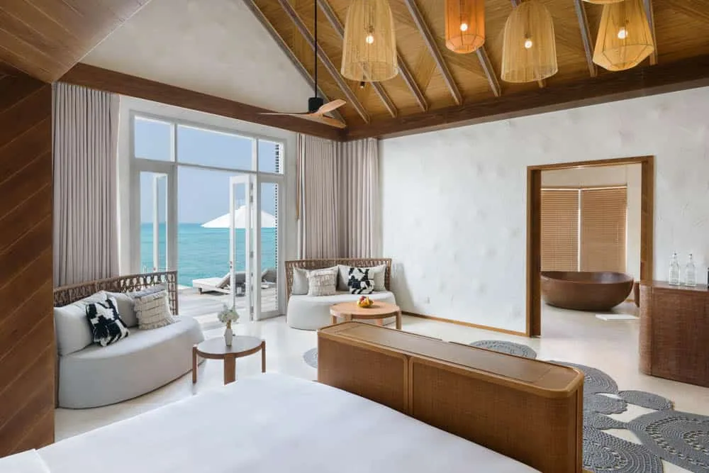 Sirru Fen Fushi Private Lagoon Resort, Water Sunset Pool Villa interior with rattan pendant cluster, dark wood beam ceiling, rattan-framed sofas and dark-wood vessel bathtub overlooking lagoon, Shaviyani Atoll, Maldives