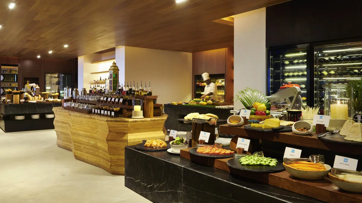 Sirru Fen Fushi Private Lagoon Resort, Raha Market all-day venue with chef at live cooking station, walnut paneling, charcuterie and cheese on stone tile and wine cooler, Shaviyani Atoll, Maldives
