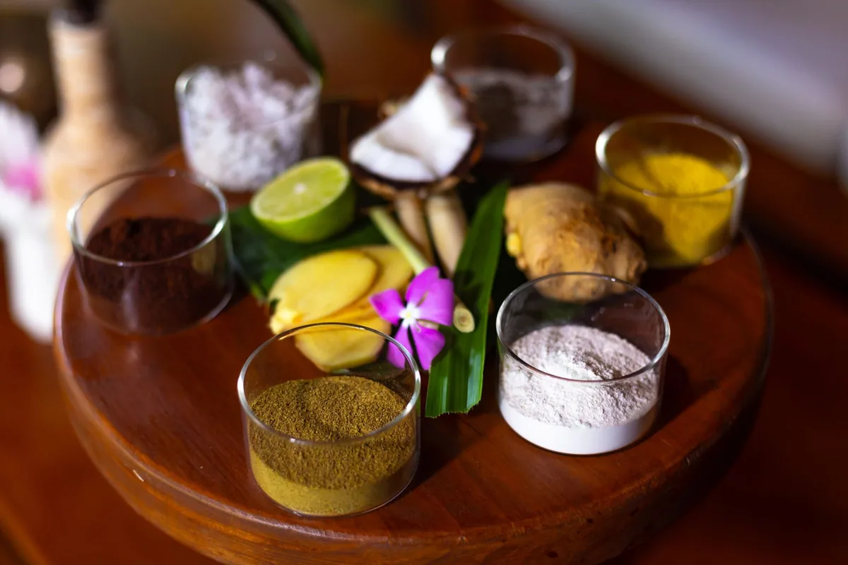 Fihalhohi Island Resort, spa, South Malé Atoll, Maldives, Spa ingredient preparation tray: glass cups holding ground coffee, kaolin clay, salt, and turmeric powder arranged around fresh coconut half, lime, ginger root, mango slices, and pandan leaves on a polished wooden tray, single pink orchid as accent, the on-property wellness programme at the budget-leaning treatment depth.