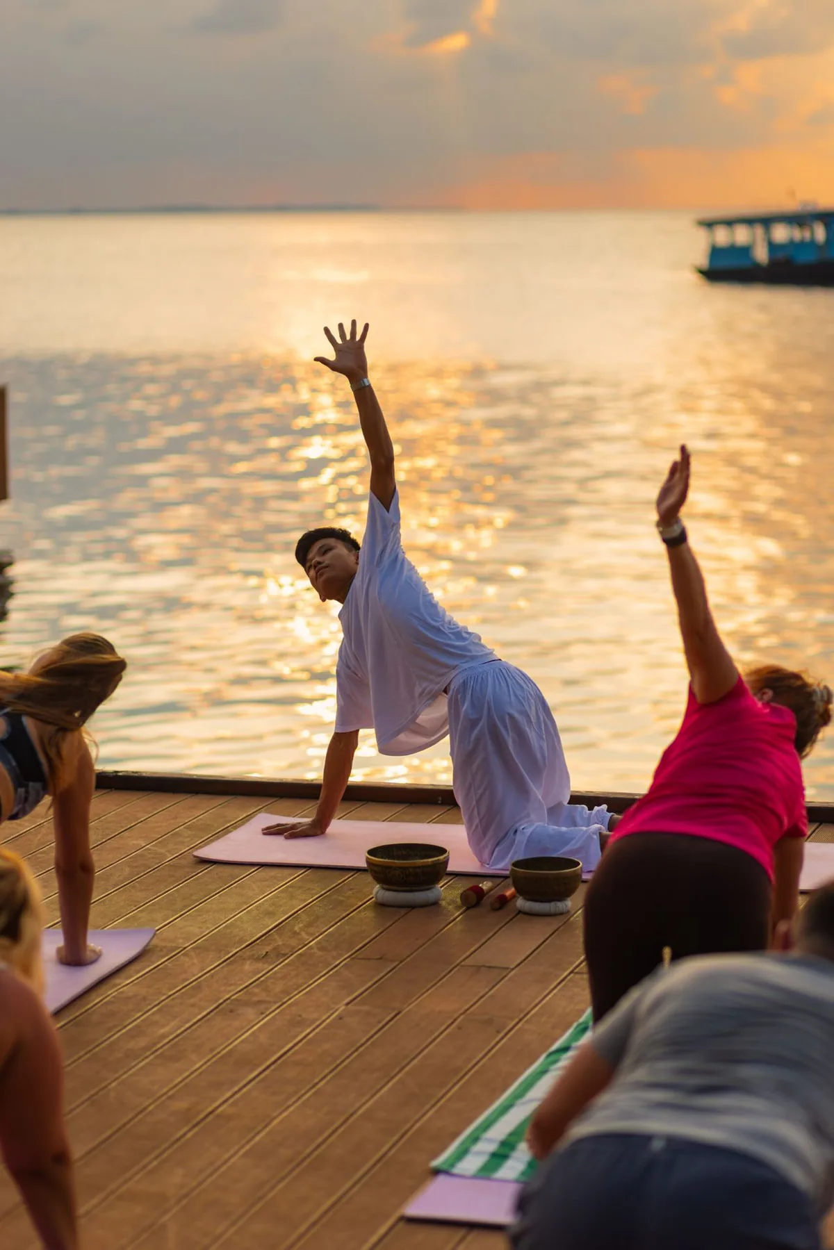 Fihalhohi Island Resort, wellness, South Malé Atoll, Maldives, Sunset yoga session on the overwater wooden deck: instructor in white leading a side-stretch pose, students mirroring on mats with brass singing bowls between them, the property's signature blue dive boat anchored on the water beyond, the lagoon catching the post-sunset gold reflection.