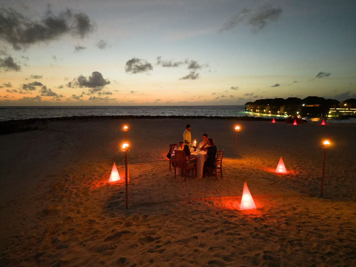 Fihalhohi Island Resort, dining, South Malé Atoll, Maldives, Private beach dinner at twilight: candle-lit table for two on white sand, conical red lanterns ringing the setup, the overwater villa cluster glowing in the distance, the post-sunset sky shading pink to deep blue (Fihalhohi's signature romantic beach-dining programme).