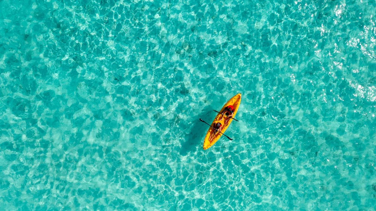 Fihalhohi Island Resort, activity, South Malé Atoll, Maldives, Top-down drone aerial of a single yellow kayak with two paddlers gliding across the property's shallow turquoise house-reef lagoon, the clear water revealing the seafloor sand texture beneath.