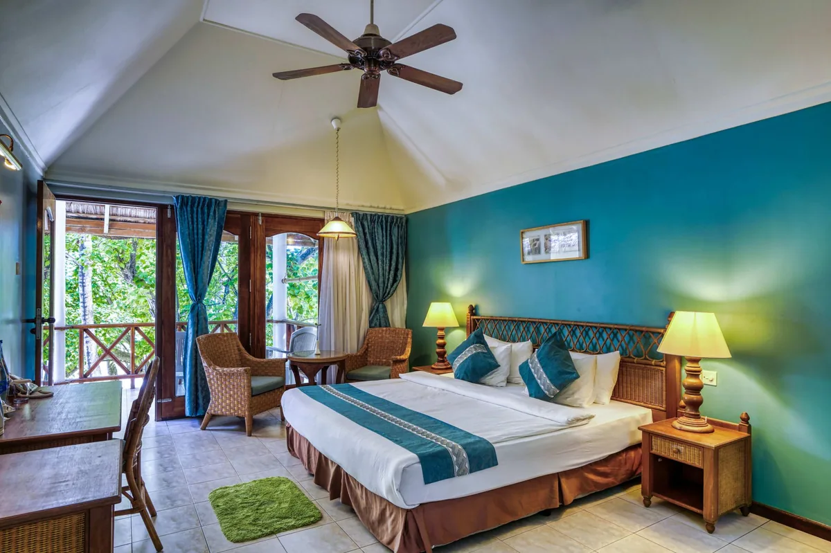 Fihalhohi Island Resort, villa beach, South Malé Atoll, Maldives, Villa interior at the traditional Deluxe Sky tier: peaked white ceiling with wooden ceiling fan, teal textured accent wall, wicker headboard, twin lamps on turned-wood nightstands, French doors opening to a warm-wood X-balustrade balcony framed by green foliage, the older, traditional villa template.