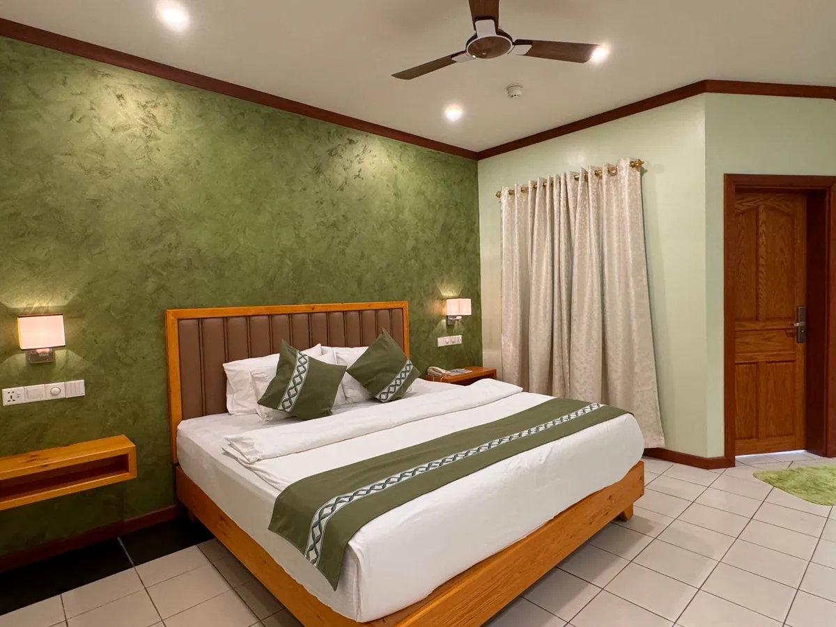 Fihalhohi Island Resort, villa beach, South Malé Atoll, Maldives, Villa interior at the renovated contemporary tier: flat white ceiling with single-paddle fan, modern leatherette panelled headboard, green textured accent wall, green-and-white runner across white linen, light-tile floor, the post-renovation contemporary villa template alongside the heritage configurations.