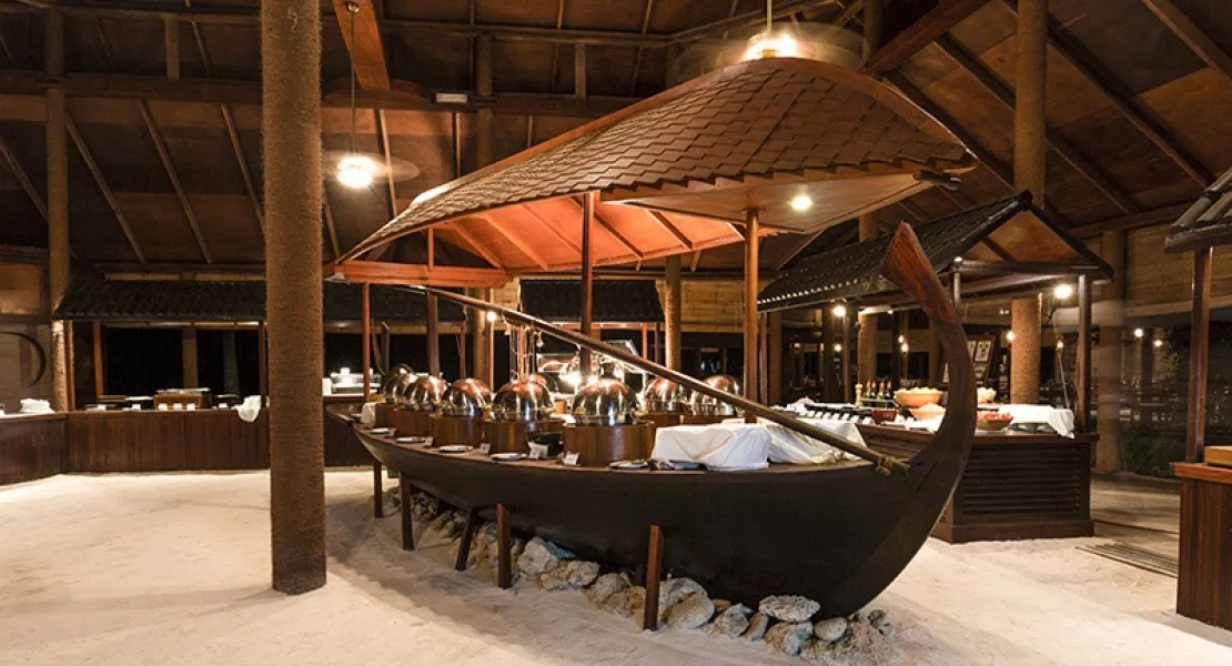 Filitheyo Island Resort, dining, Faafu Atoll, Maldives, Main restaurant interior with the signature traditional Maldivian dhoni-boat-as-buffet display station, the dhoni's curved hull is the centerpiece of the open pavilion with copper-domed chafing dishes lined along the gunwale, sand floor and dark-timber columns supporting the thatched ceiling.