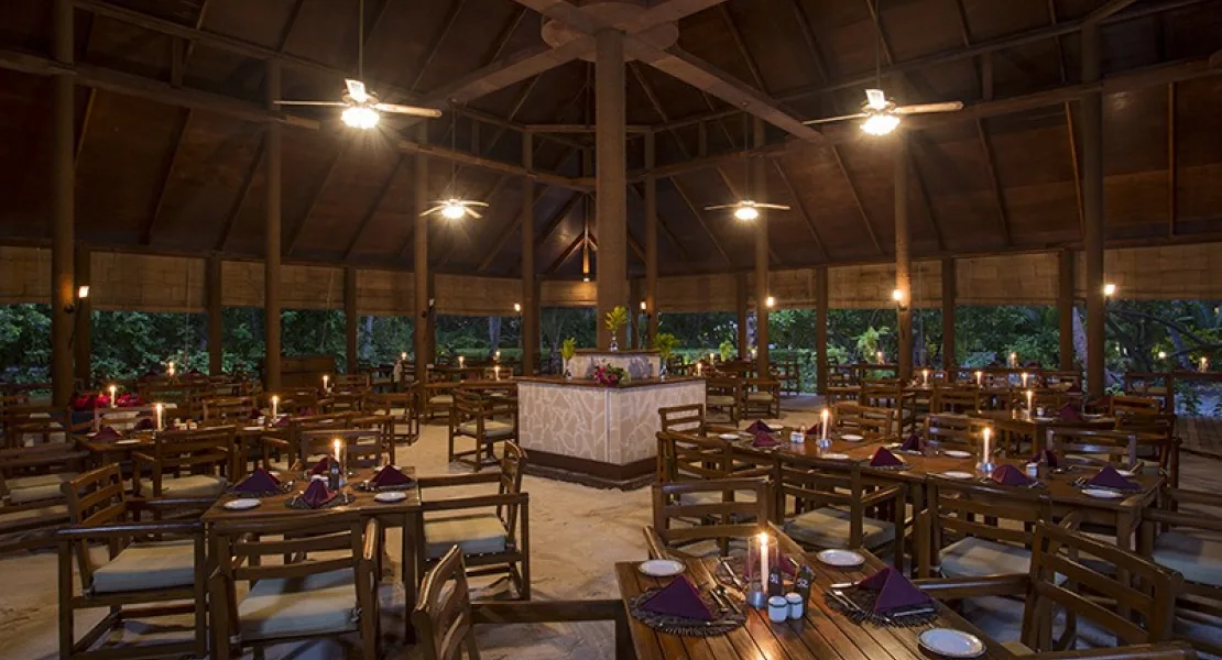 Filitheyo Island Resort, dining, Faafu Atoll, Maldives, Main restaurant dining hall at night, vaulted timber-beam pavilion with a central column, dark-timber tables set with red napkins and lit candles on the sand floor, ceiling fans overhead and the jungle visible at the open perimeter.