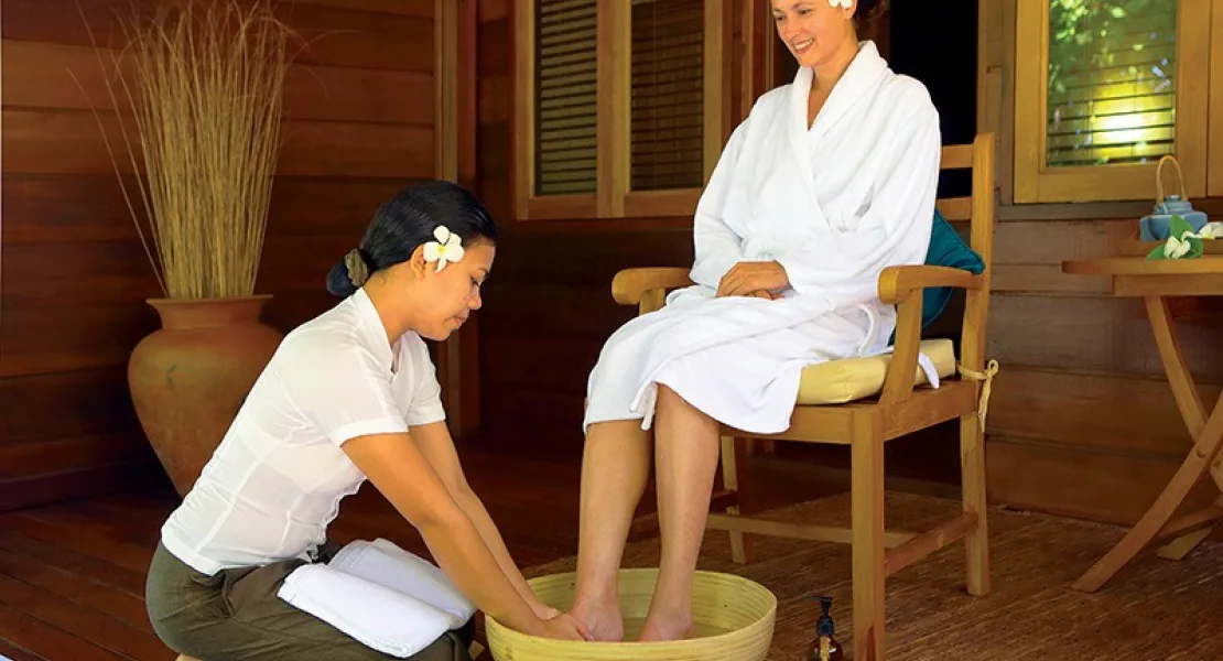Filitheyo Island Resort, spa, Faafu Atoll, Maldives, Spa welcome ritual on the dark-timber pavilion veranda, a therapist with plumeria washing a guest's feet in a wooden bowl, ceramic vase and tea-set on the side table at right.