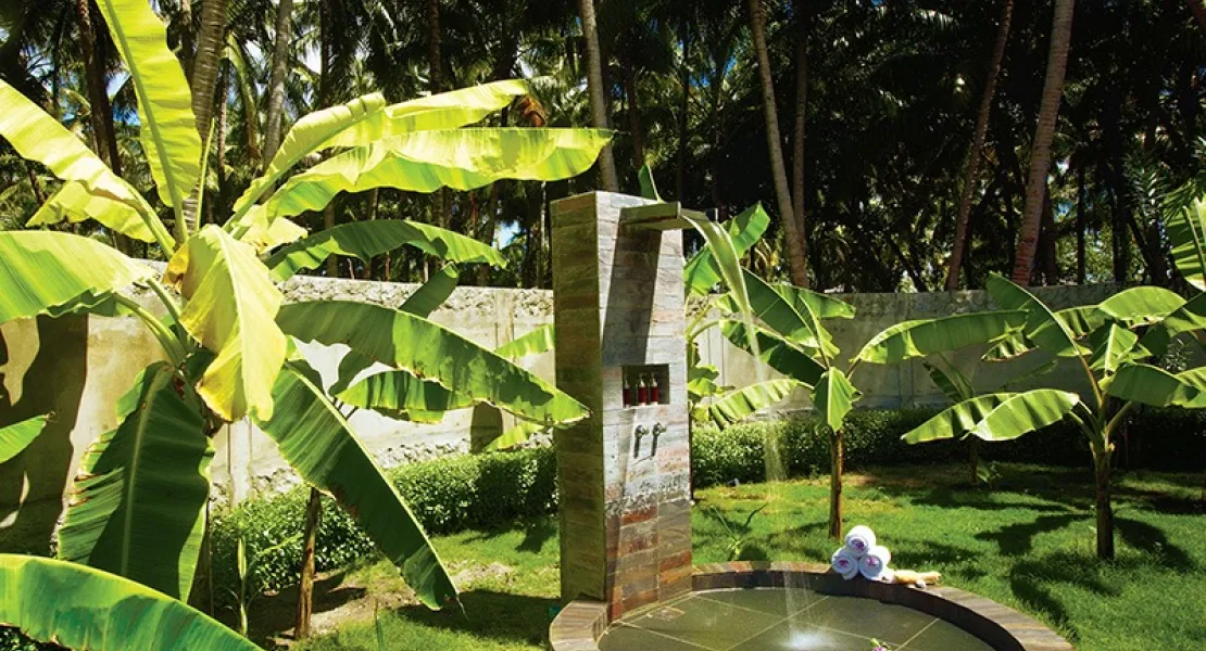 Filitheyo Island Resort, spa, Faafu Atoll, Maldives, Outdoor spa shower garden, a slate-clad shower pillar and a circular timber-rim plunge basin with steaming water, banana-tree garden enclosed by concrete walls, palm-canopy backdrop.