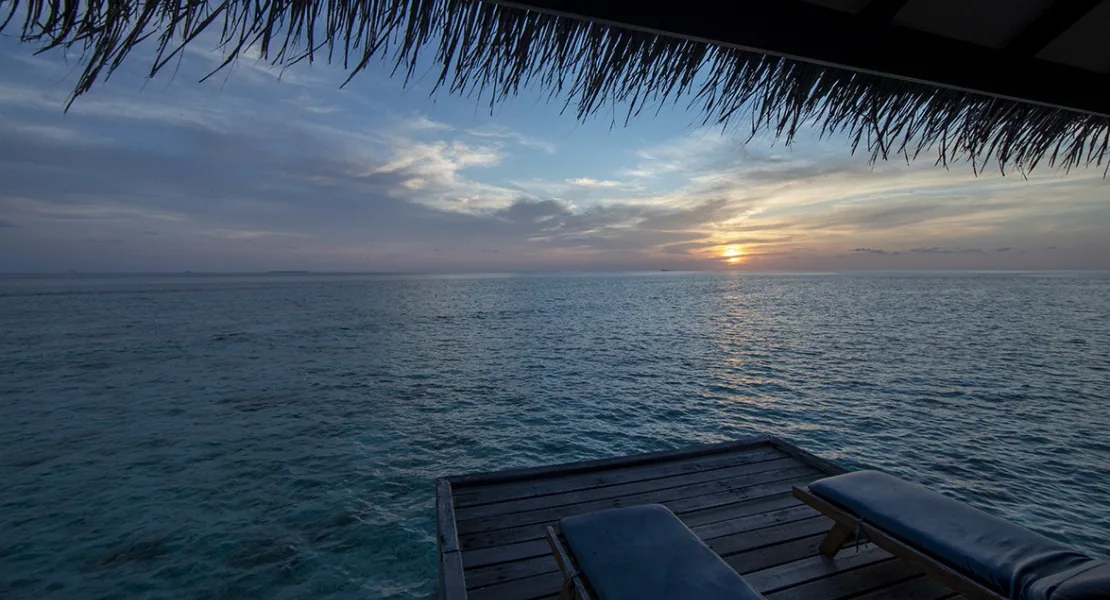 Filitheyo Island Resort, villa overwater, Faafu Atoll, Maldives, Water Villa deck at sunset, thatched eave fringing the top of the frame, dark-timber sun loungers on the deck, calm lagoon stretching to a sun-setting horizon.