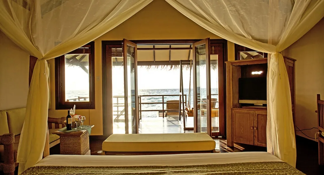 Filitheyo Island Resort, villa interior, Faafu Atoll, Maldives, Water Villa bedroom interior, four-poster bed with sheer mosquito-net canopy, daybed cushion, dark-timber cabinetry with TV, open doors leading onto the over-water deck with a chair and the lagoon beyond.