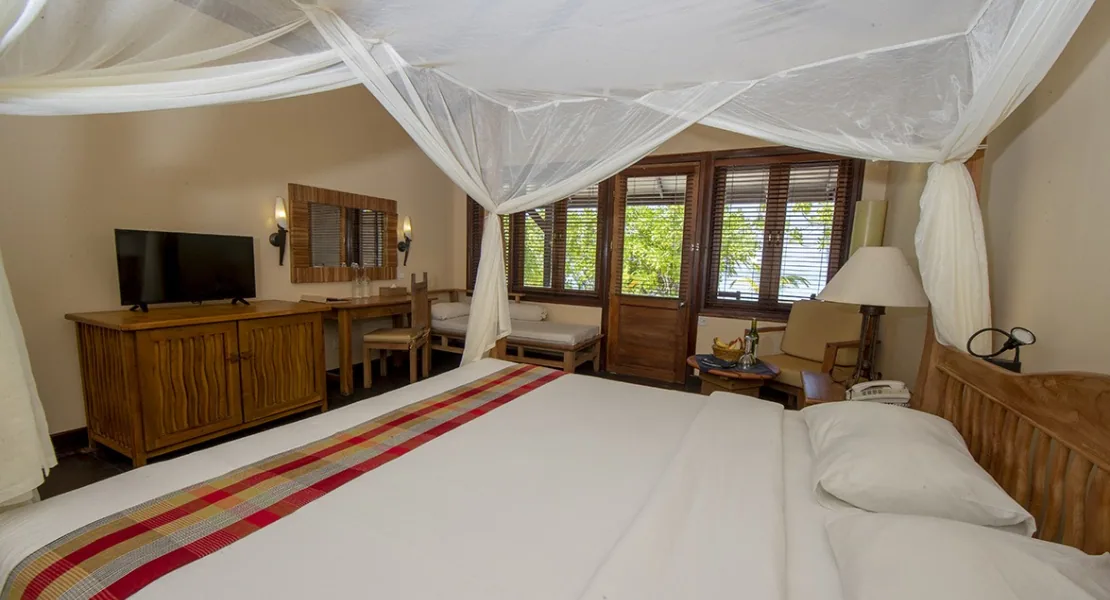 Filitheyo Island Resort, villa interior, Faafu Atoll, Maldives, Superior Villa bedroom, four-poster bed with sheer canopy and red-and-yellow striped bed runner, dark-wood cabinetry with TV, wooden louvered windows, dining table at the side.