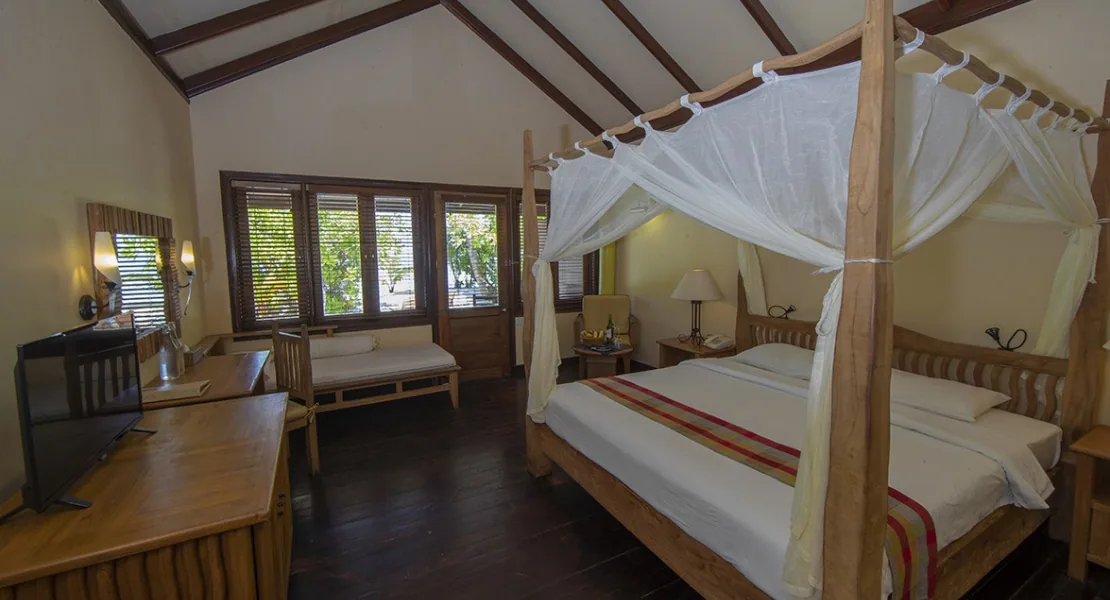 Filitheyo Island Resort, villa interior, Faafu Atoll, Maldives, Superior Villa wide interior, vaulted timber-beam ceiling, four-poster bed with sheer net canopy at right, daybed at left, dark-hardwood floor and wooden louvered windows.