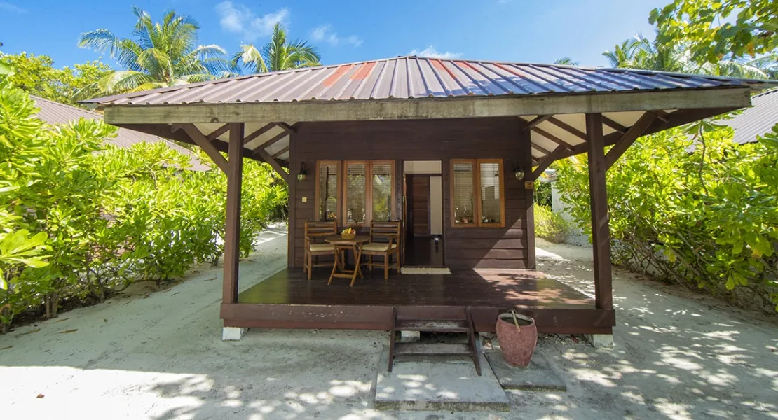 Filitheyo Island Resort, villa beach, Faafu Atoll, Maldives, Deluxe Villa exterior, dark-stained timber villa with corrugated metal roof and covered verandah with wooden table and chairs, sand walkway through palm canopy.