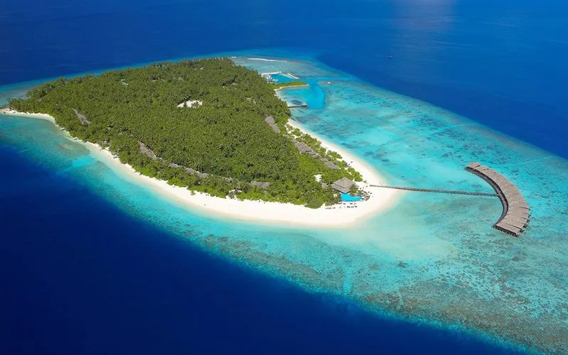 Filitheyo Island Resort, aerial, Faafu Atoll, Maldives, Filitheyo Island Resort full-island drone aerial: oval palm-canopy island geometry, white sand beach perimeter, central pool deck, and the long over-water villa jetty extending from the right reef edge into the lagoon, central Faafu Atoll.