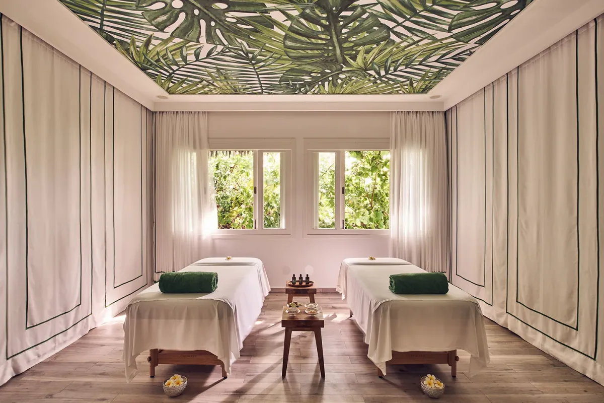 Finolhu Baa Atoll, spa, Baa Atoll, Maldives, Cowrie Spa twin treatment room interior, the paired teak-frame massage beds with white linens and folded green hand-towels, brass tray of treatment oils on the central low-table, palm-leaf-print ceiling installation above and the garden glimpsed through the cream-curtained windows.