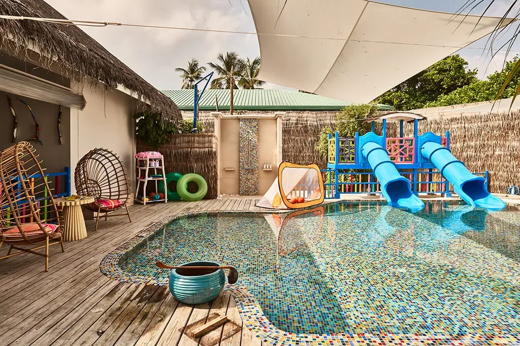 Finolhu Baa Atoll, activities, Baa Atoll, Maldives, Oceaneers Kids Club pool with the curved blue twin-tube waterslide arching into the mosaic-tile splash pool (the tile floor laid in the property kaleidoscope-colour pattern), teal-yellow-and-pink rattan loungers on the surrounding deck under the white shade-sail canopy.