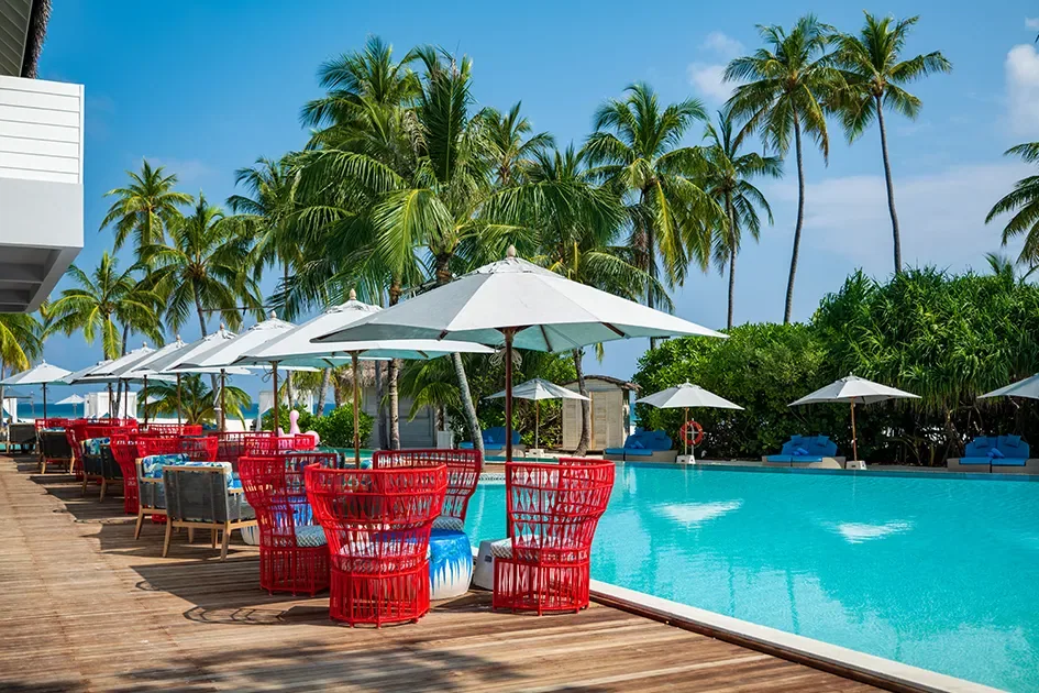 Finolhu Baa Atoll, dining, Baa Atoll, Maldives, Beach Club pool deck, the row of red and cobalt-blue cane-back chairs lining the long pool bar, cream parasols casting hard shadows on the hardwood decking, tall coconut palms framing the lagoon view.