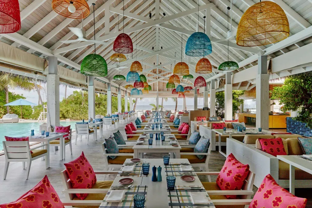 Finolhu Baa Atoll, dining, Baa Atoll, Maldives, Beach Kitchen all-day pavilion interior, the signature kaleidoscope-colour cane pendant lights suspended over the long communal table, pink and cobalt and teal cushioned woven chairs lining the bench seating and the open-eave white pavilion opening to the lagoon.